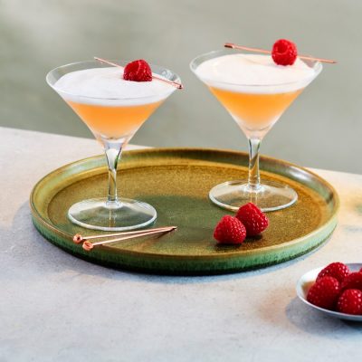 Two frothy pink French Martinis garnished with fresh raspberries placed on a silver serving platter on a table covered in a white tablecloth with a bowl of fresh raspberries and one more French Martini in the foreground