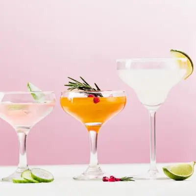 Front view of five cocktails in different glasses against a pale pink backdrop