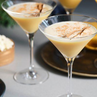 A trio of indulgent Eggnog Martini cocktails presented in long-stemmed Martini glasses, surrounded by snack bowls on a light grey surface