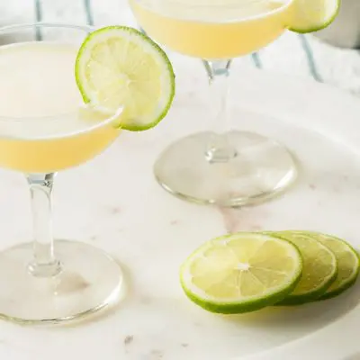 Close up top view of two Gimlet Cocktails in coupe glasses, garnished with lime wheels, and presented on a white platter set on a striped blue and white tablecloth