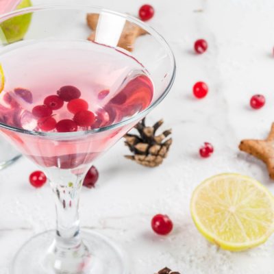A pretty Cranberry Martini garnished with fresh cranberries, against a white backdrop studded with pine cones and ginger biscuits in the shape of stars