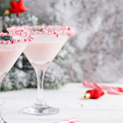 Two Peppermint Martinis with candy rims against a snowy backdrop with Christmas decorations to the side