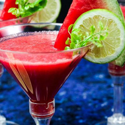 Two Watermelon Martinis garnished with a lemon wedge and slice of watermelon each, against a blue backdrop