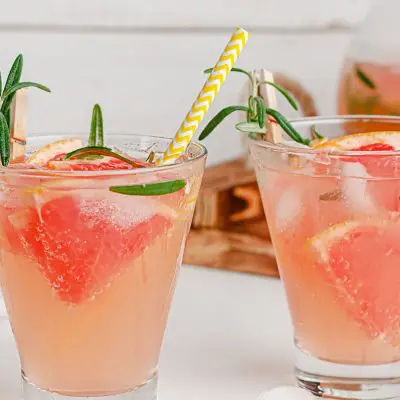 Close up of a pair of Greyhound Cocktails garnished with fresh grapefruit, on a white surface, with sliced grapefruit and a jug of juice in the background