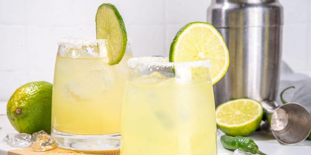 A marvellous Mezcal Margarita with lime and a salt rim against a white backdrop with a cocktail shaker and lime wedge to the side