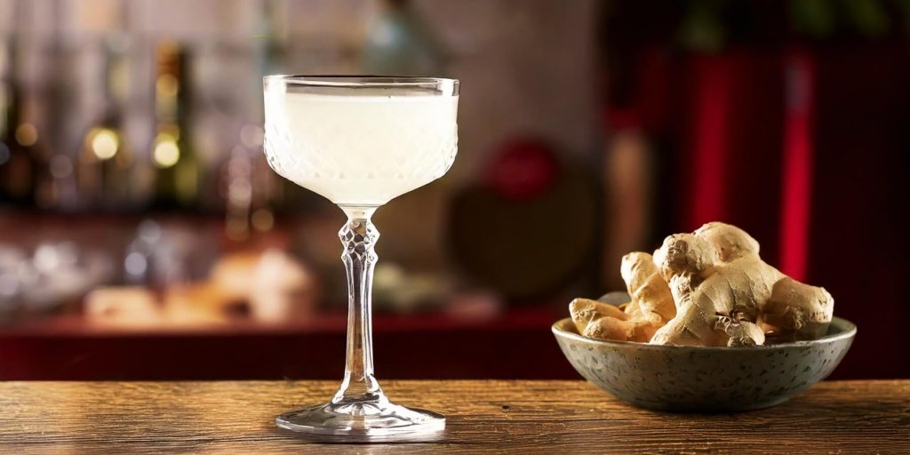 An elegant coupe glass filled with a pale, ginger-infused cocktail, placed next to a bowl of fresh ginger roots on a rustic wooden table in a cozy bar setting.