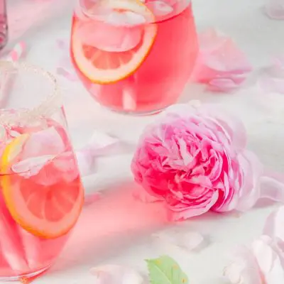 Three pretty pink Mother's Day cocktails against a white backdrop with a pink rose and some petals scattered for a touch of whimsy