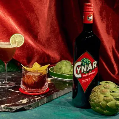 A sumptuous collection of retro lush cocktails posed next to a bottle of Cynar liqueur agains a blue marble backdrop draped with red velvet