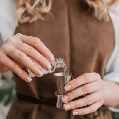 Close up front shot of a woman in a blonde woman in a brown apron pouring clear liquid into a metal jigger from a small bottle