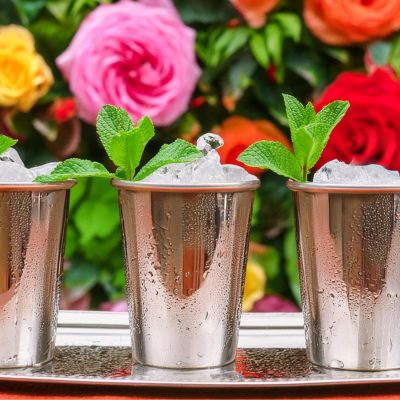 Three Mint Julep Kentucky Derby cocktails served in pewter cups, vertical rose garden in the background