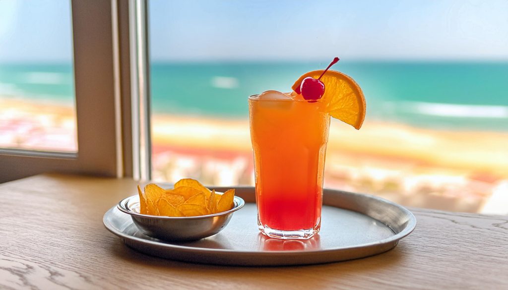 A SKYY Vodka Sex on the Beach cocktail served on a tray with a bowl of crisps, ocean view through open window in background