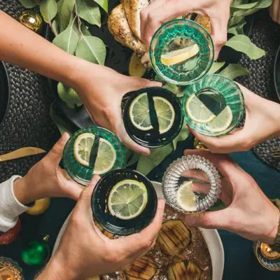 Top view of a group of friends clinking together festive Christmas cocktails over a table filled with holiday foods