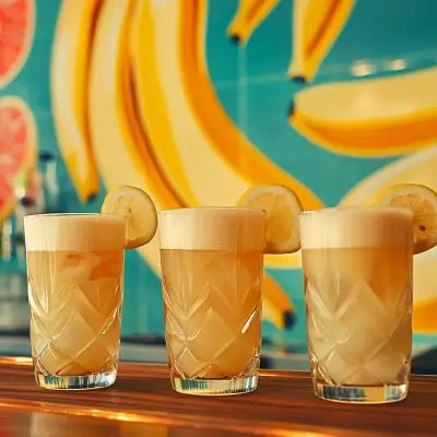 Three banana liqueur cocktails on a wooden bar counter against a wall covered in bold banana theme wallpaper