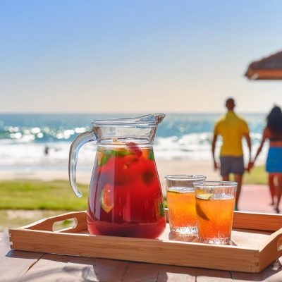 A tray with a jug of summer rum cocktails and two glasses on a wooden table outside a modern holiday home with a beach scene in the background