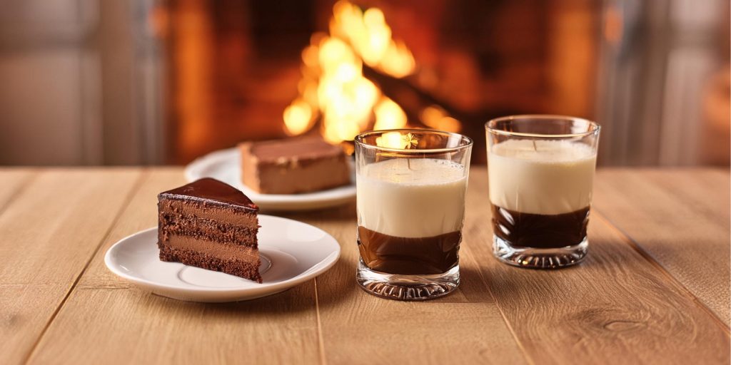 Two White Russian cocktails on a wooden table next to serving plates with portions of chocolate cake, with a cosy fire in the background