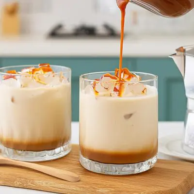 Close up of caramel sauce being poured into one of two Salted Caramel White Russians in a modern kitchen setting