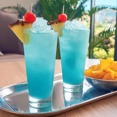 Two Frozen Blue Hawaiian cocktails with pineapple wedge and cherry garnish, served with a bowl of crisps