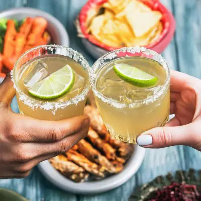 Two hands clinking together Reposado Tequila Margarita cocktails over a Mexican themed lunch table