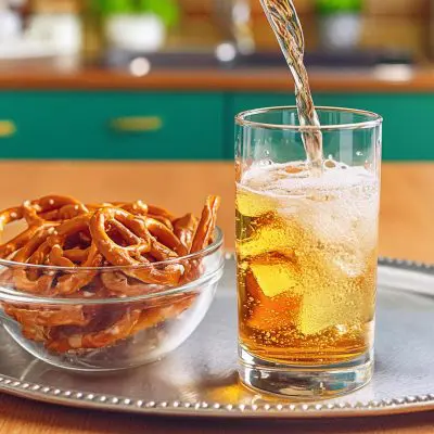 A mixed bourbon drink being poured into a tall glass on a silver platter next to a bowl of pretzels