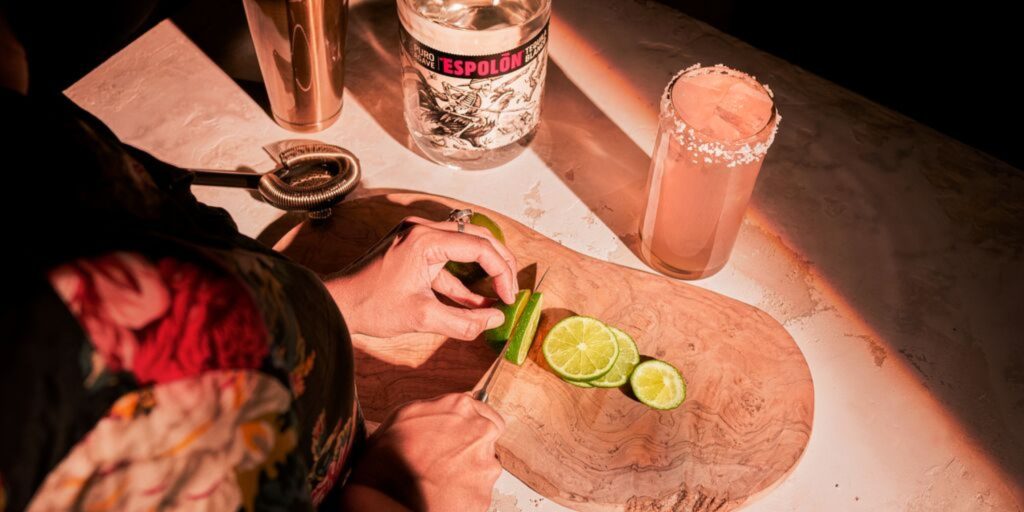 Overhead shot of a bartender cutting lime slices with a pink Paloma cocktail to the side