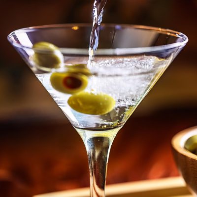 A stream of vodka being poured into a martini glass with green olives