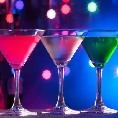 Three brightly coloured 90s cocktails served in a club setting with neon lights in the background