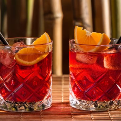 Two bright red Kingston Negronis with orange garnish, bamboo in the background