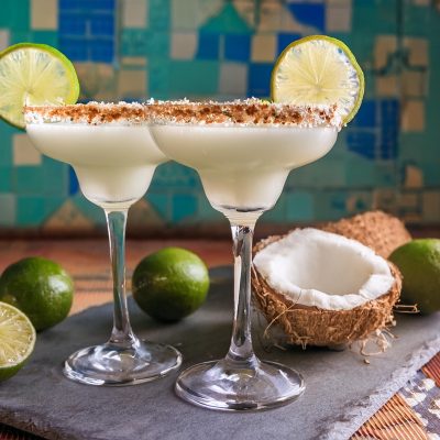 Two Coconut Margarita cocktails on a table in a modern home lounge against a blue patterend backdrop, surrounded by fresh coconuts and limes