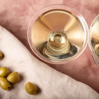 An overhead shot of a gin martini and a vodka martini on a dusty pink surface with olives to the side