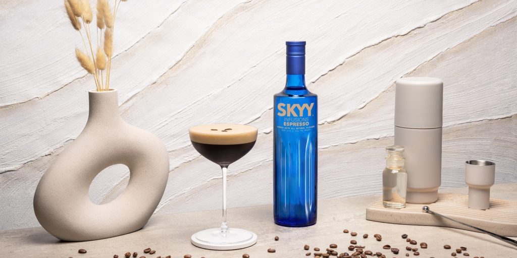 An Espresso Martini with a foamy top, garnished with three coffee beans, next to a blue bottle of SKYY Infusions Espresso, cream and white themed background and props