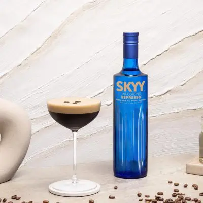 An Espresso Martini with a foamy top, garnished with three coffee beans, next to a blue bottle of SKYY Infusions Espresso, cream and white themed background and props