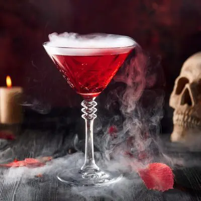 Close-up of a deep red smoking Vampire's Kiss vodka cocktail, candles and skull in the backgound