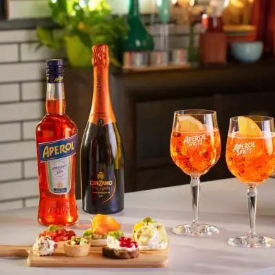 A styled shot of two vibrant orange Aperol Spritz cocktails next to a bottle of Aperol and Cinzano Prosecco