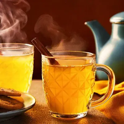 Two streaming Espolòn Tequila Hot Toddy cocktails in glass mugs with cinnamon garnish and blue kettle in the background