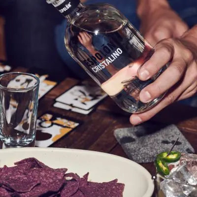 A close-up shot of a hand that's about to pour Espolòn Cristalino tequila into a glass on a table with playing cards and beetroot chiips