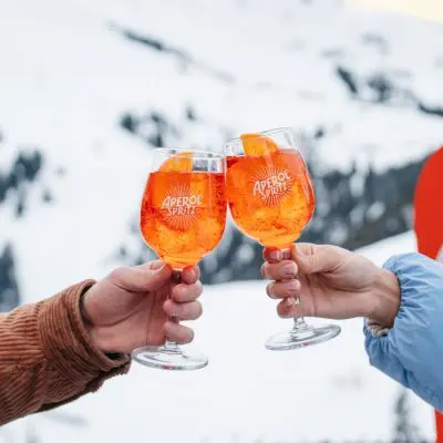 Two hands clinking together bright orange Aperol cocktails in an Apres Ski setting, snowboard and snowy mountains in the background