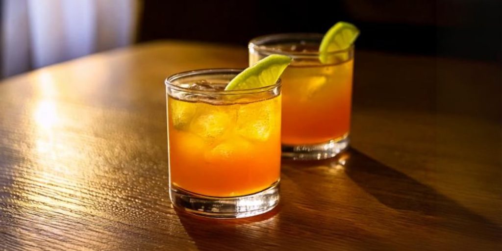 Two amber-hued Naked in the Desert cocktails in lowball glasses with ice cubes, garnished with lime wedges, set on a warmly lit wooden surface.