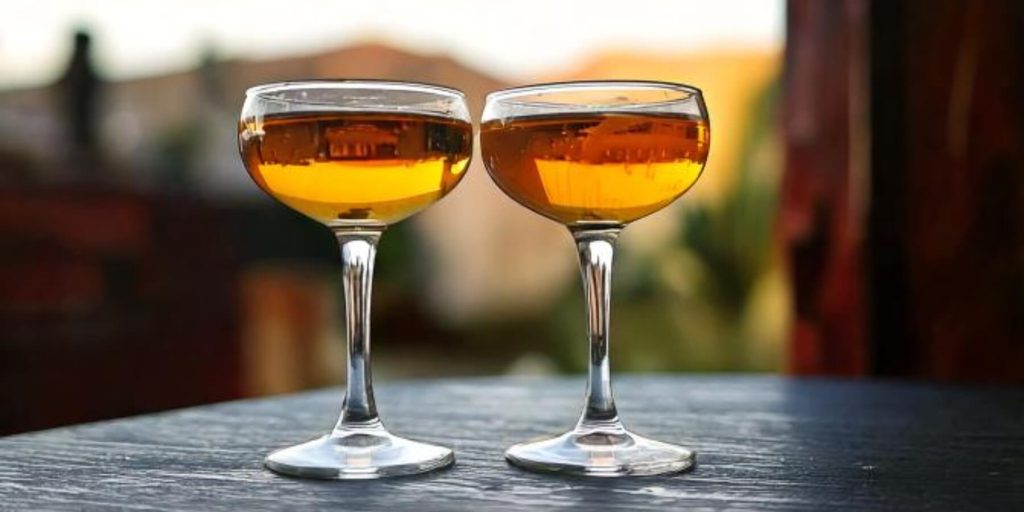 Two golden Round 2 cocktails served in coupe glasses, placed on a dark outdoor table with a blurred sunset view in the background.