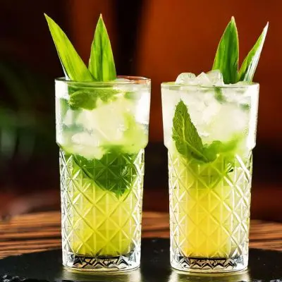 Two refreshing green Mojitos garnished with mint leaves and pandan leaves, served in tall crystal glasses filled with ice, on a wooden table.