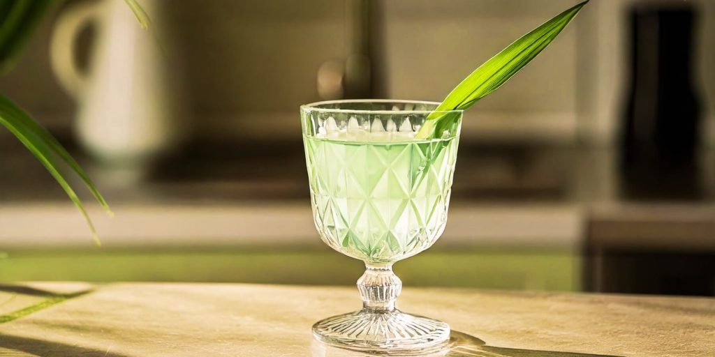 A pale green cocktail served in an elegant, vintage-style glass, garnished with a single pandan leaf, with a soft kitchen background.