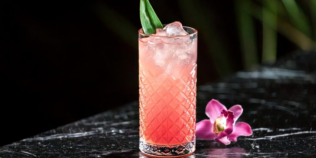 A vibrant pink cocktail garnished with a fresh green pandan leaf, served in a tall crystal highball glass with ice, accompanied by a delicate purple orchid.