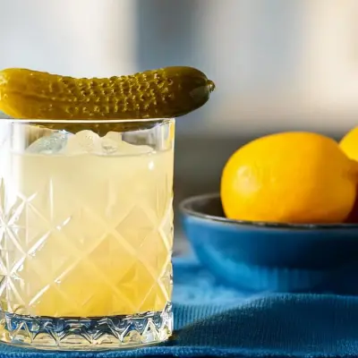 A short glass with a pale yellow cocktail over ice is garnished with a whole pickle resting on the rim. Behind the drink, a blue bowl holds fresh lemons, complementing the cocktail's bright citrus notes.