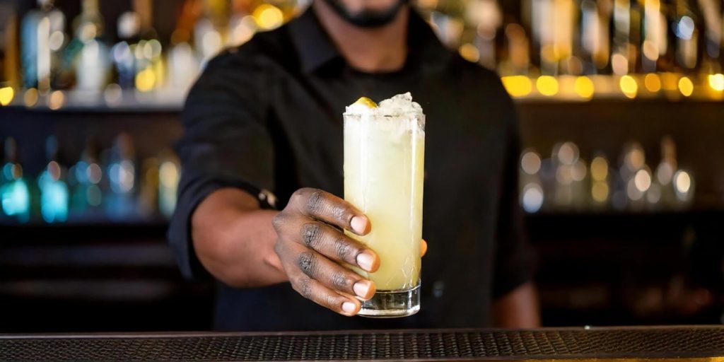 A bartender, dressed in black, presents a tall glass filled with an ice-cold yuzu cocktail. The blurred background of a bar with golden lighting creates a stylish, inviting atmosphere.