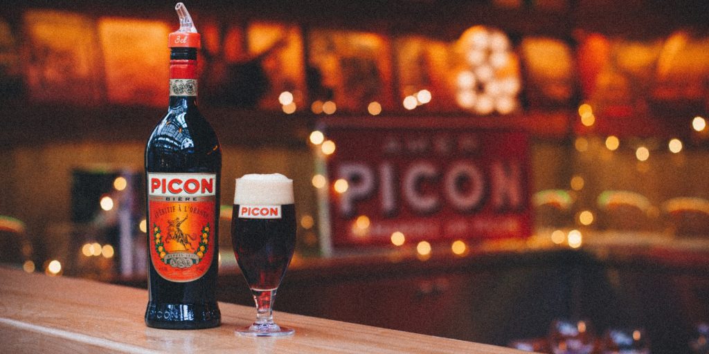 A Picon Biére cocktail on a bar counter with a Picon sign in the background