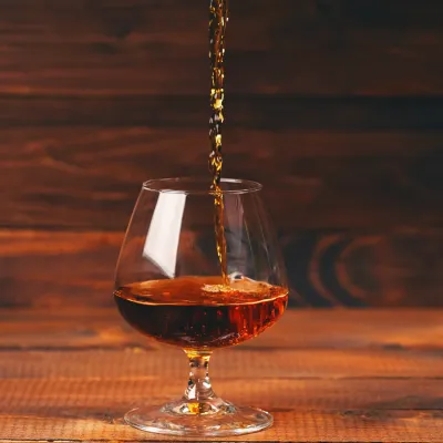 Pouring shot of cognac into a glass on a wooden table, photo by levgenii_Meyer