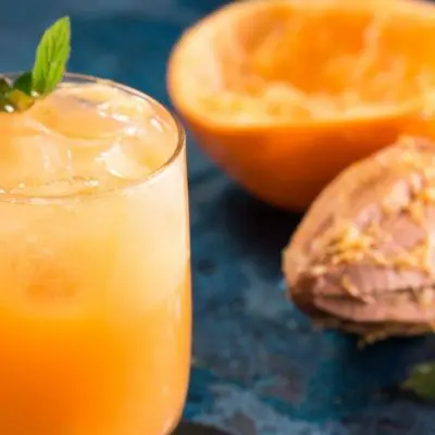 Top view of an Orange Blossom cocktail in a tumbler, garnished with a sprig of mint, featured on a blue surface with an orange in the background