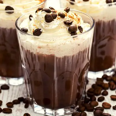 Close-up of a trio of decadent Mexican Coffee Cocktails with whipped cream topping, garnished with coffee beans, posed on a white surface scattered with coffee beans