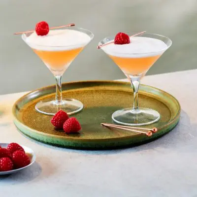 Two frothy pink French Martinis garnished with fresh raspberries placed on a silver serving platter on a table covered in a white tablecloth with a bowl of fresh raspberries and one more French Martini in the foreground