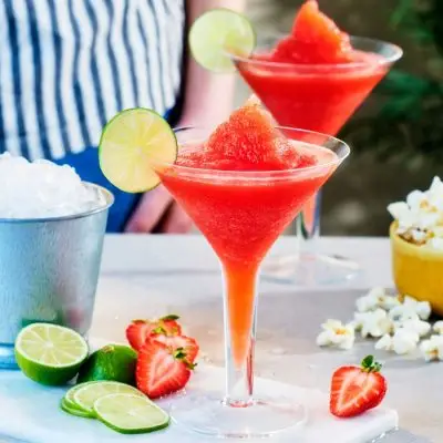 Front view of two glasses of Frozen Strawberry Daiquiri on a white table, surrounded by snacks, with a person in a blue striped shirt in the background