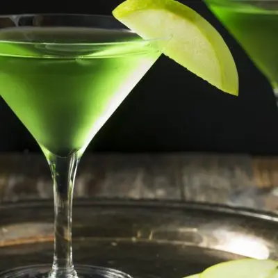 Close up of two bright green Apple Martinis with sliced apple garnish, presented on a small silver tray, set on a rustic wooden surface